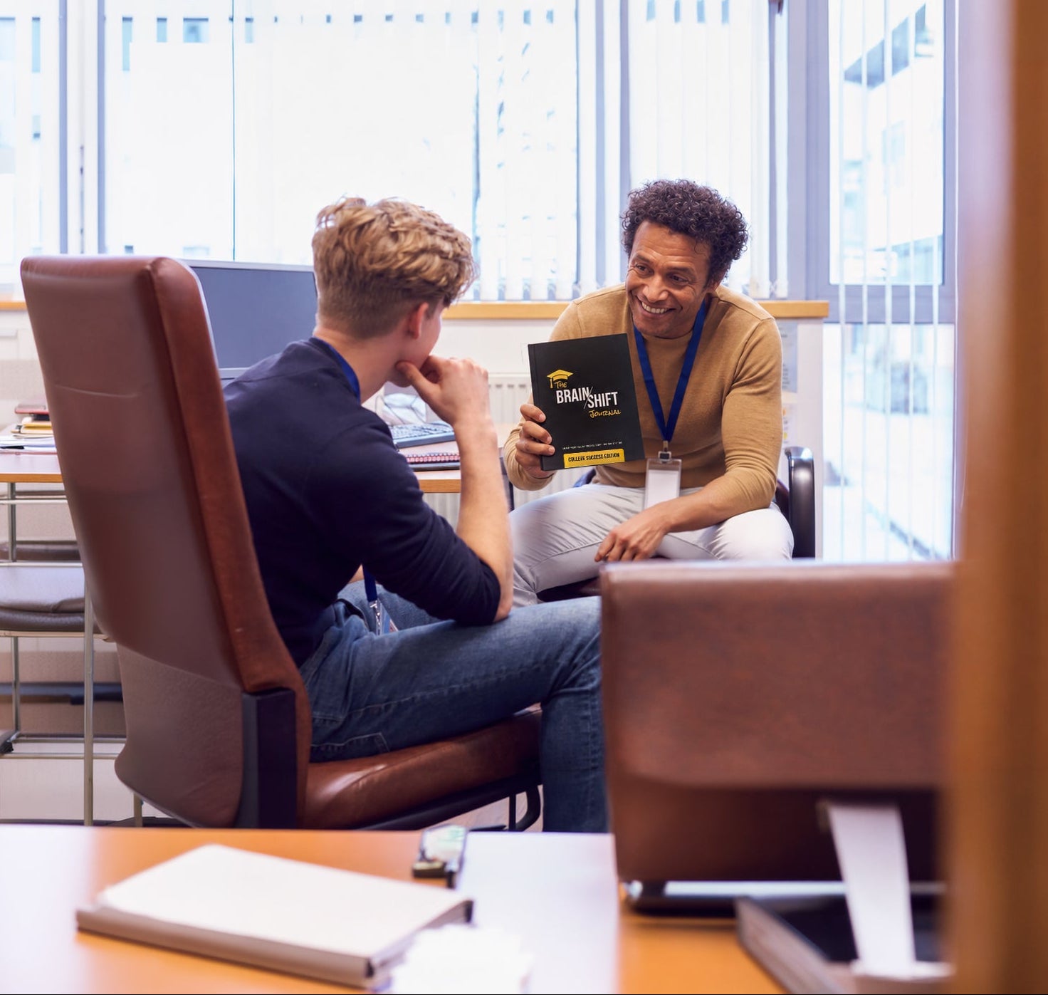 Two people sitting at a table with a laptop and a book in an office setting.