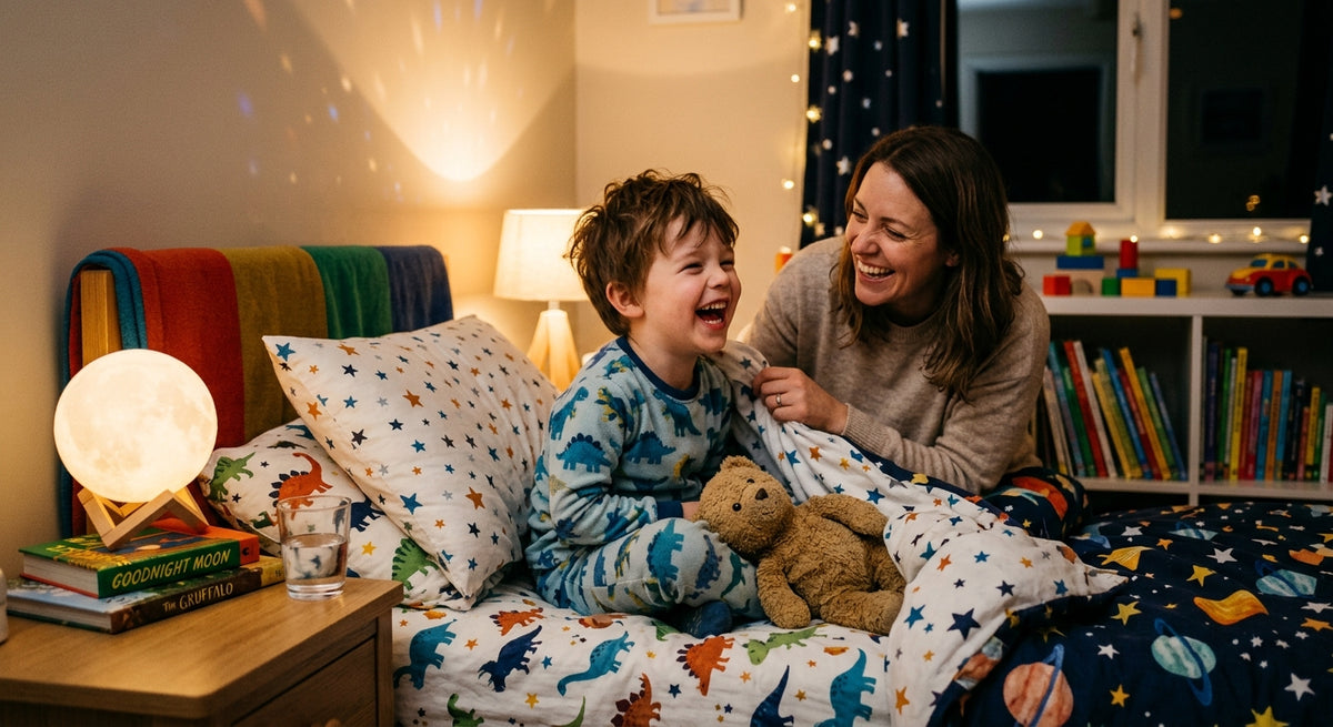 A cozy children's bedroom scene with a young boy in dinosaur-print pajamas sitting in bed laughing with a woman, surrounded by matching dinosaur and space-themed bedding, a moon lamp, and books on a shelf.